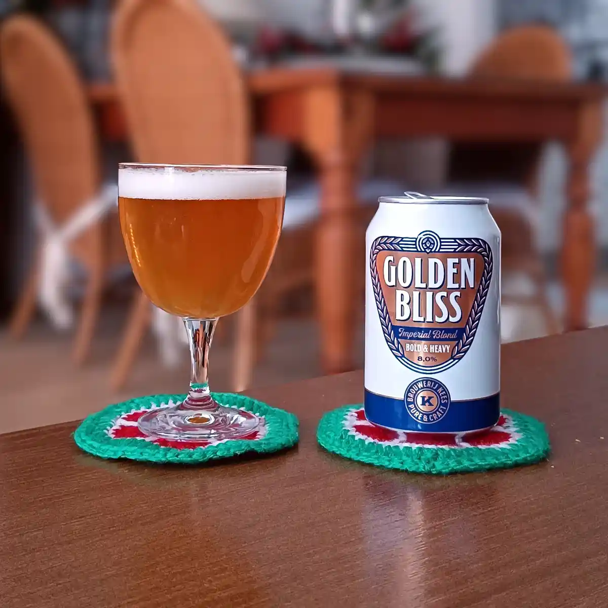 A can of 'Golden Bliss' Imperial Blond beer and a glass of the same beer sit side-by-side on colorful crocheted coasters on…