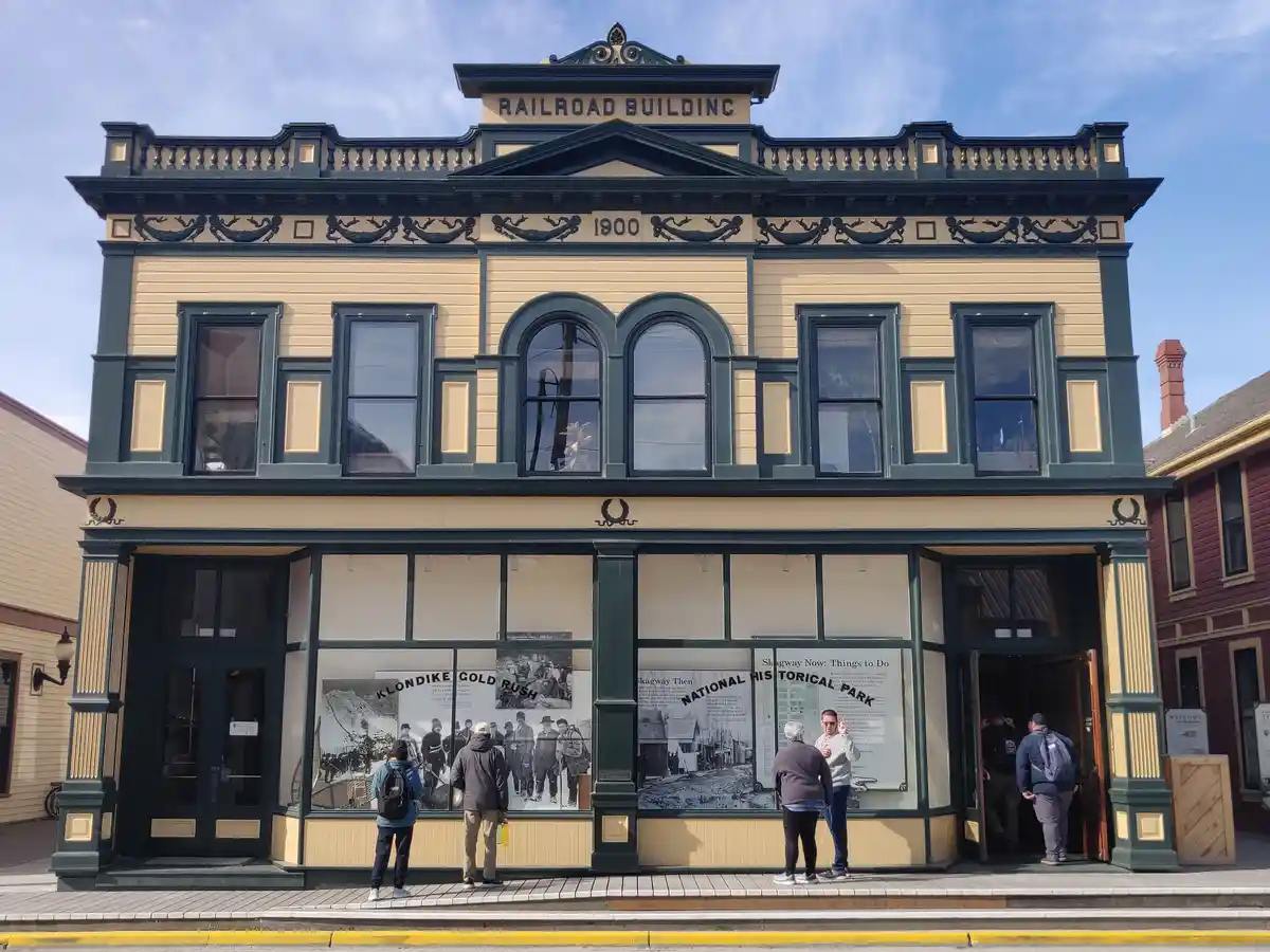 A historic two-story yellow and green building labeled 'RAILROAD BUILDING 1900' with tourists viewing historical exhibits in…