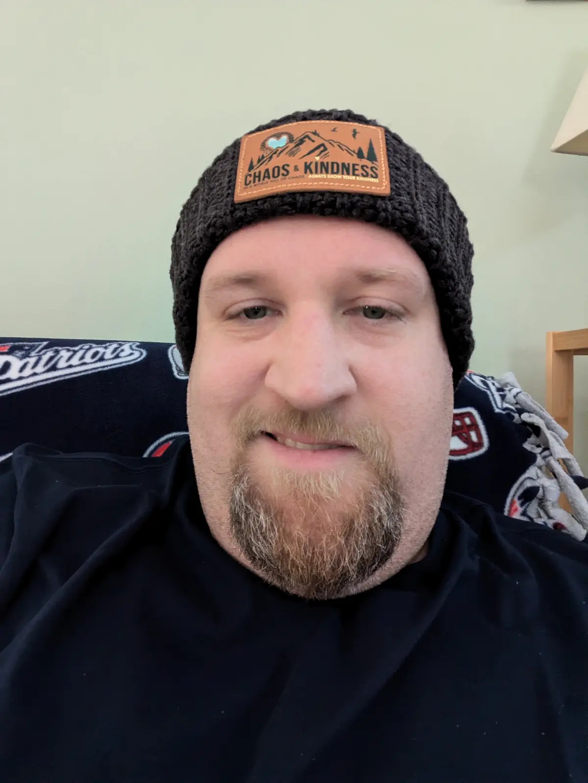 A man with a beard and mustache wearing a dark knit beanie with a brown leather patch that reads 'CHAOS & KINDNESS IN A…