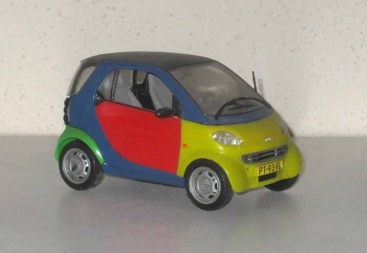 A toy model of a Smart car is shown with a multi-colored body: blue roof, red door, yellow front, and green fender. The car…