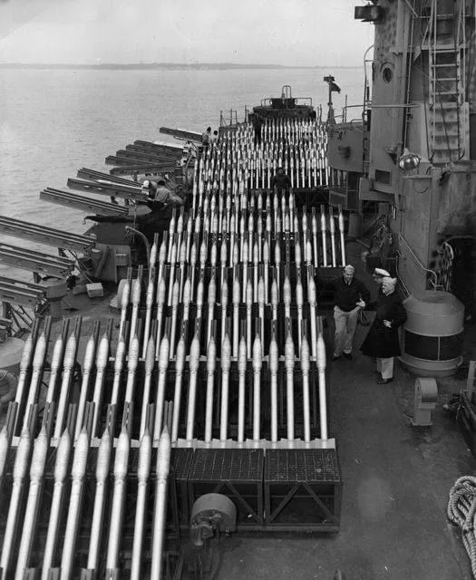 A black-and-white historical photo shows two people standing on the deck of a warship, surrounded by rows of loaded…