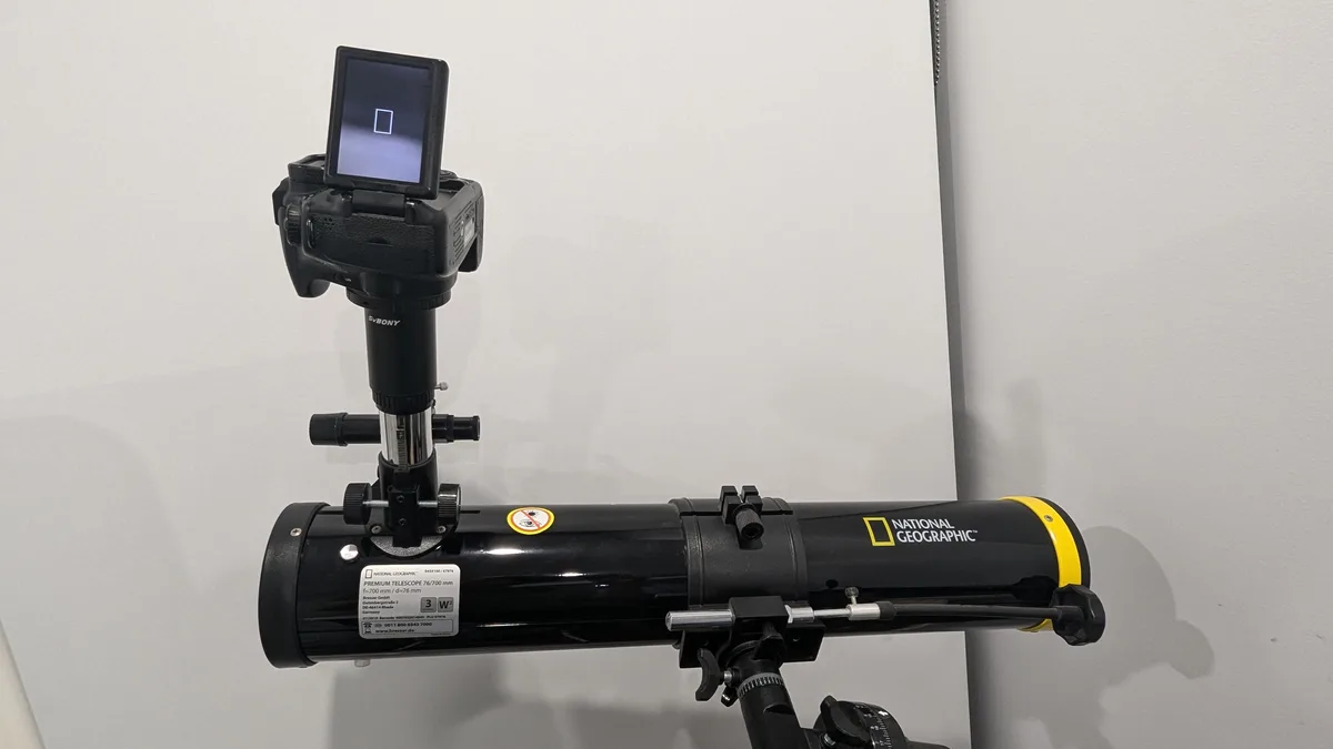 A black National Geographic branded telescope is mounted on a tripod and pointed upward, with a DSLR camera mounted on top…