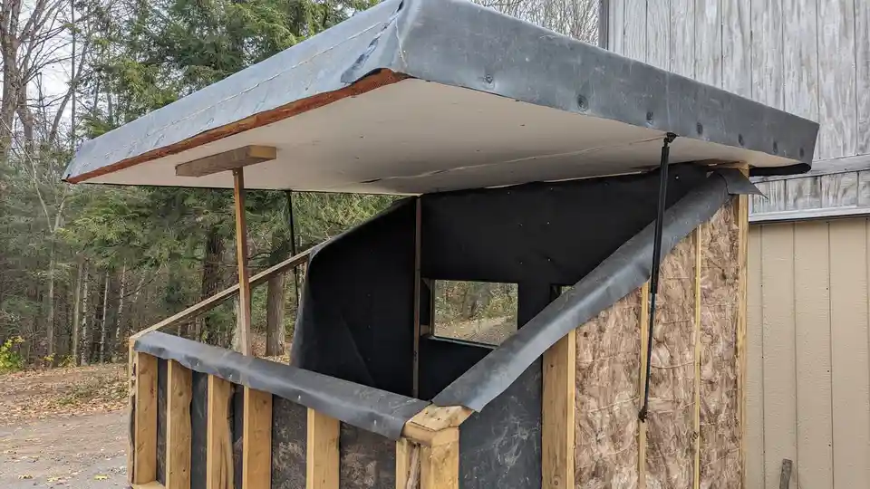 A partially constructed wooden shed with exposed insulation and black plastic sheeting, showing a makeshift roof and walls…