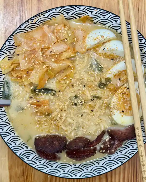 A top-down photo of a steaming bowl of ramen on a wooden table, featuring noodles, sliced boiled eggs, kimchi, sesame seeds…