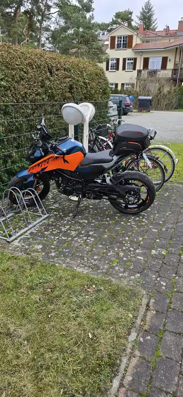A blue and orange KTM 125 motorcycle is parked on a mossy brick path next to a bike rack, with several bicycles parked…