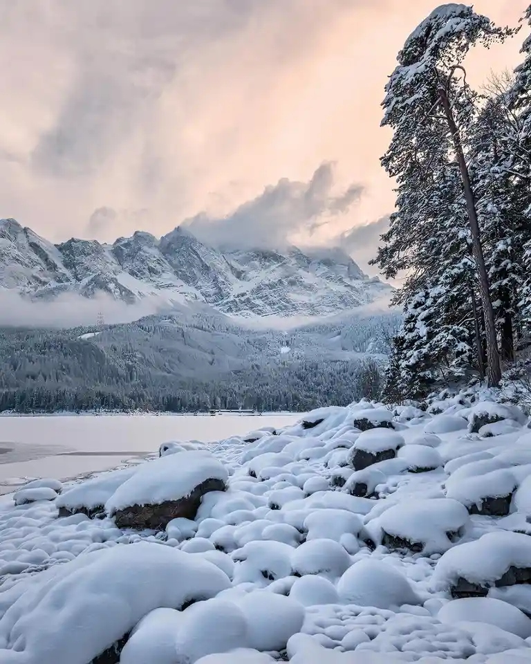 A serene winter landscape features snow-covered rocks in the foreground, evergreen trees on the right, and snow-capped…