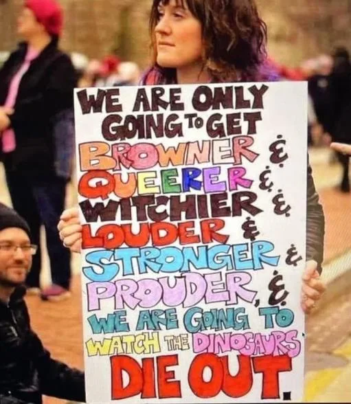 A person holds a colorful, hand-lettered protest sign with the text 'WE ARE ONLY GOING TO GET BROWNER & QUEERER & WITCHIER &…