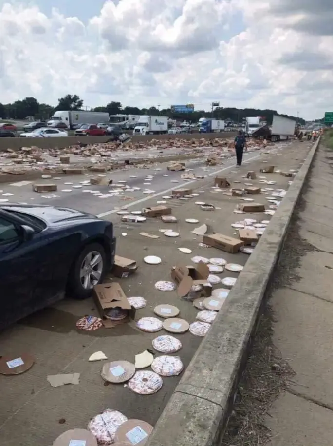A highway scene shows a black car parked on the shoulder next to a massive spill of broken pizza boxes and scattered pizza…