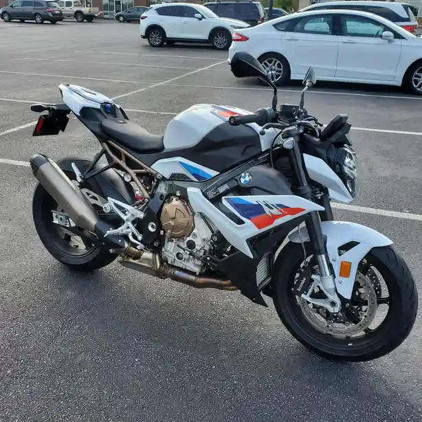 A white and black BMW S 1000 RR motorcycle is parked in a lot with cars in the background. The bike has red, white, and blue…