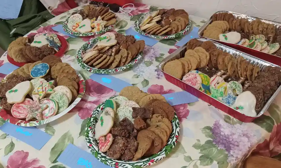 A table covered in a floral tablecloth is laden with multiple plates and trays of assorted Christmas cookies, including…