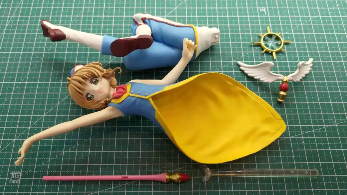 A disassembled anime figurine of a girl with brown hair, blue dress, and yellow cape lies on a green cutting mat, surrounded…