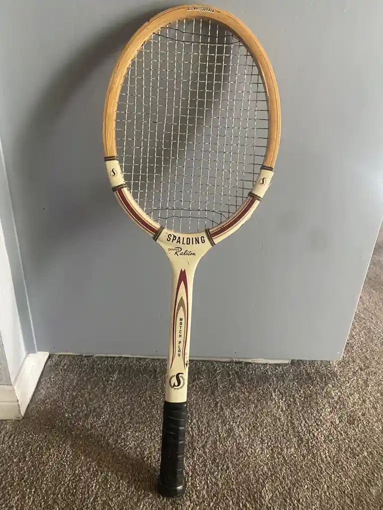 A vintage wooden Spalding tennis racket with 'Dennis Ralston' and 'Match Play' branding stands upright on a carpeted floor…