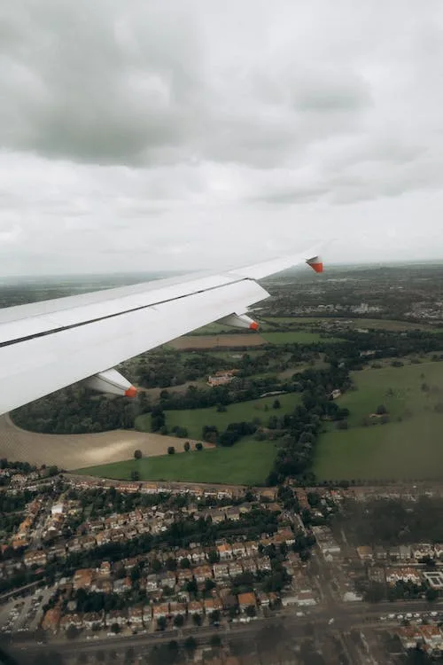 An aerial view from an airplane window shows the aircraft's wing extending over a patchwork of green fields, trees, and…