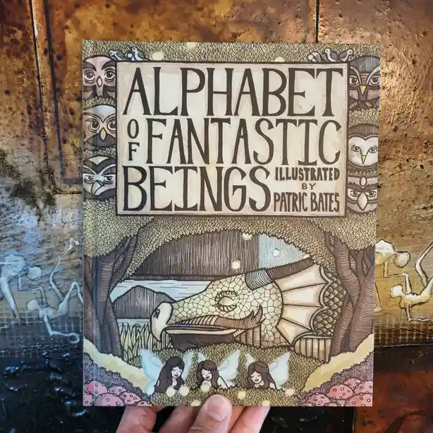 A hand holds a children's book titled 'Alphabet of Fantastic Beings' by Patric Bates, featuring a whimsical illustration of…