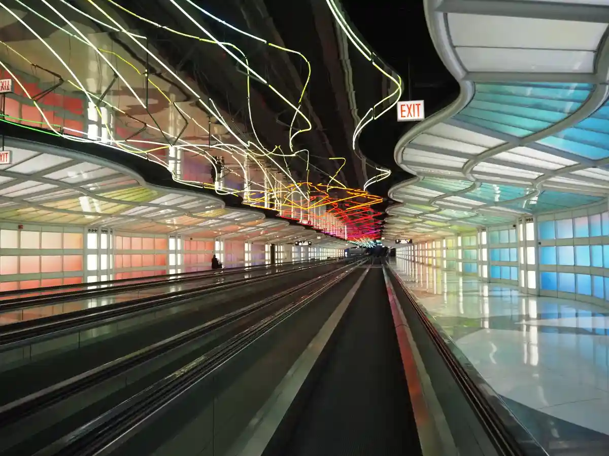 A long, empty airport moving walkway with colorful, curved ceiling lights and 'EXIT' signs. A lone person sits on the left…