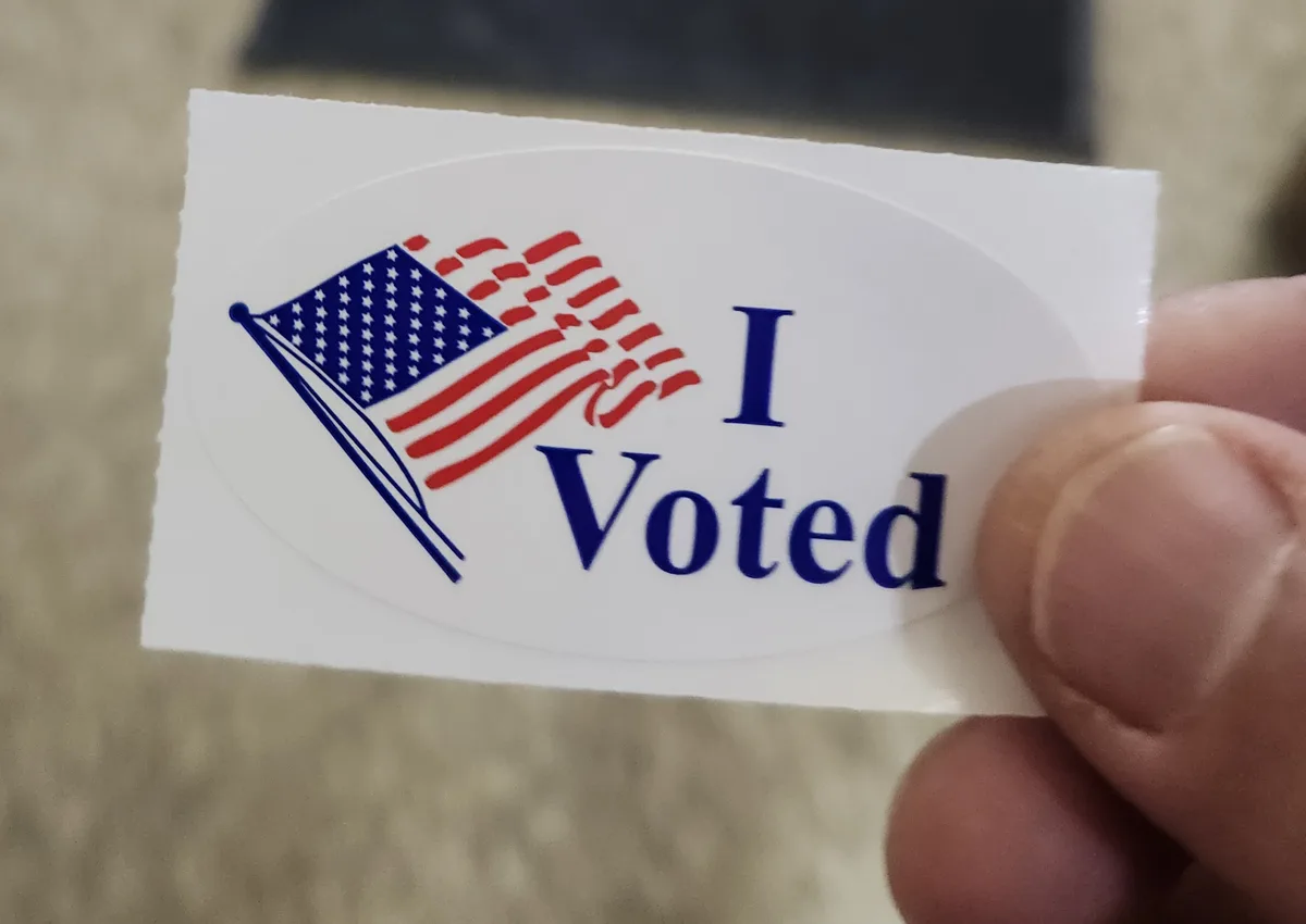 A close-up photo shows a hand holding a white oval sticker with a stylized American flag on the left and the text 'I Voted'…