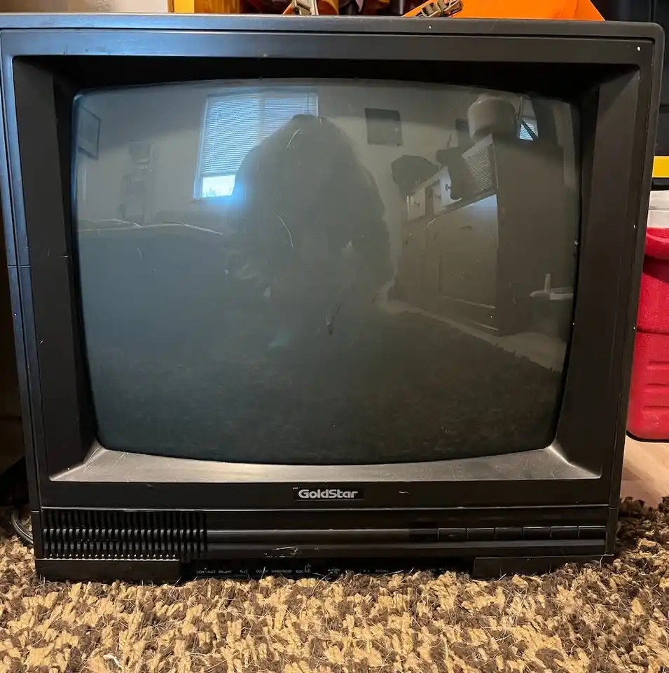 A black, boxy, old-style GoldStar CRT television sits on a textured rug, its screen dark and reflective, showing a blurry…