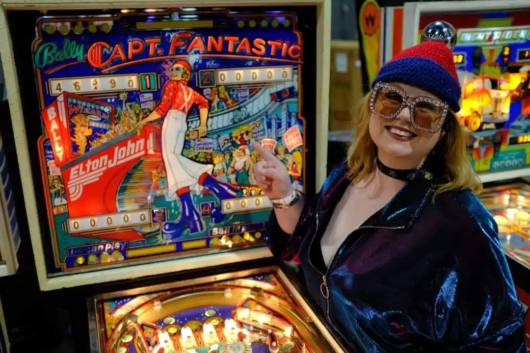A woman in a sparkly blue jacket, a red and blue knit hat with a silver ball, and large rhinestone sunglasses points at a…