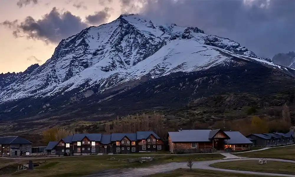 A row of rustic wooden cabins with lit windows sits in a valley at dusk, with a massive, snow-capped mountain looming behind…