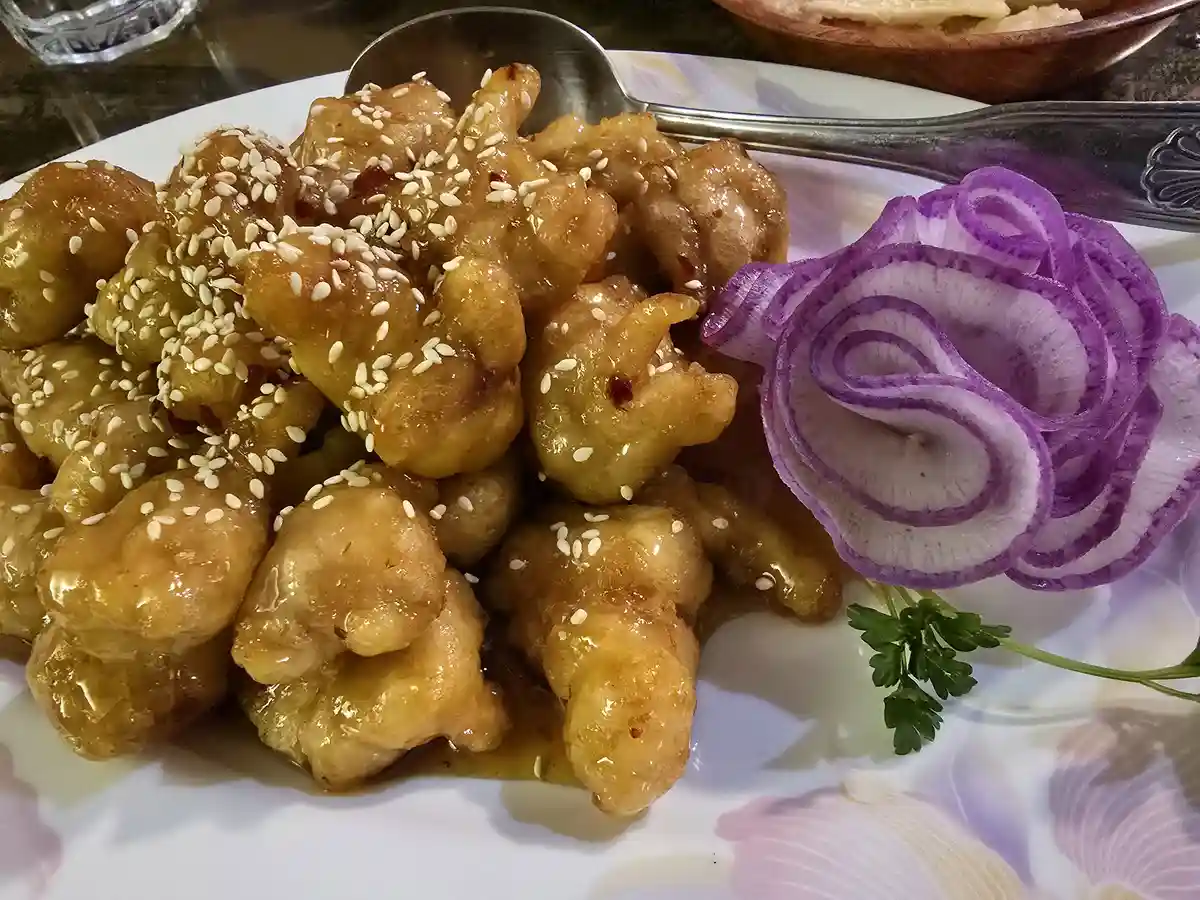 A close-up photo of a plate of sesame chicken, coated in a glossy sauce and sprinkled with white sesame seeds, garnished…