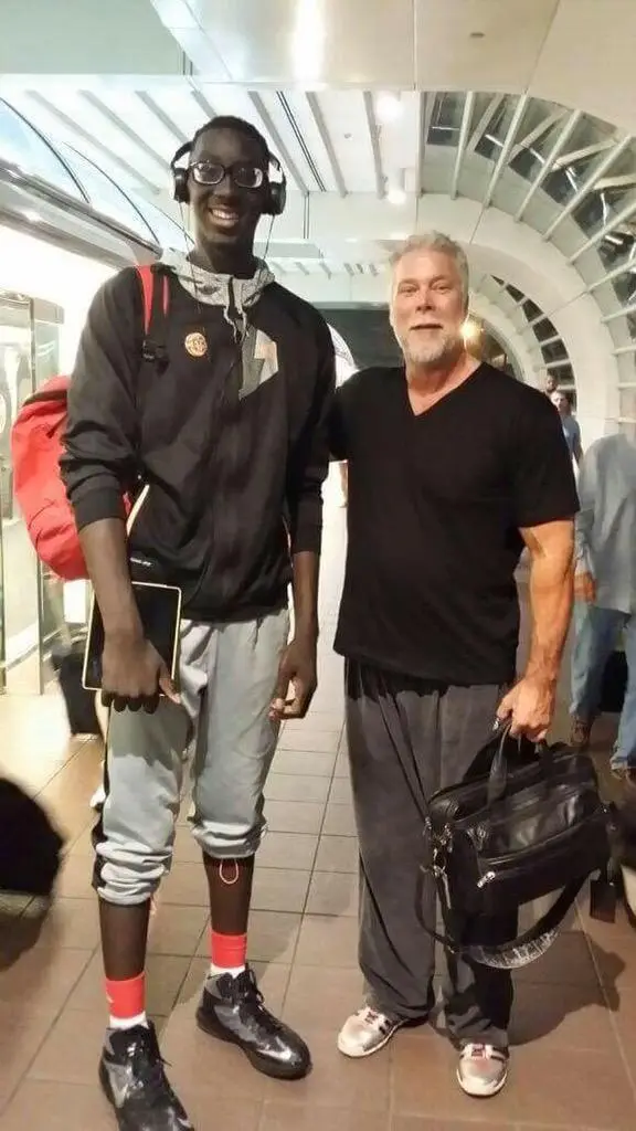 Two men stand side-by-side in an airport terminal. The man on the left is visibly much taller, wearing headphones, a black…