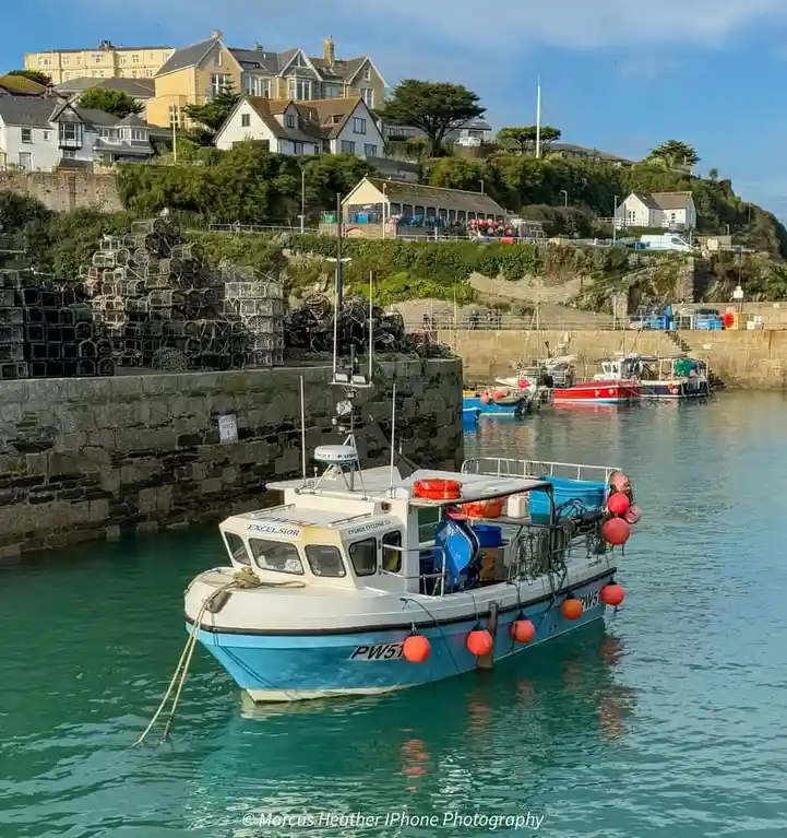 A blue and white fishing boat named 'EXCELSIOR' with registration 'PW51' is moored in a harbor, surrounded by stacks of…