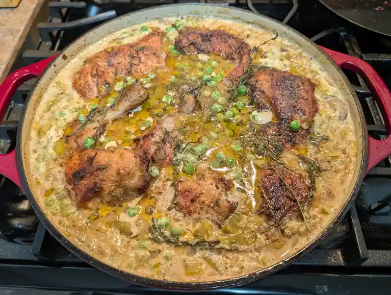 A red cast-iron skillet on a stovetop contains chicken pieces in a creamy sauce with peas and thyme. The dish appears rich…