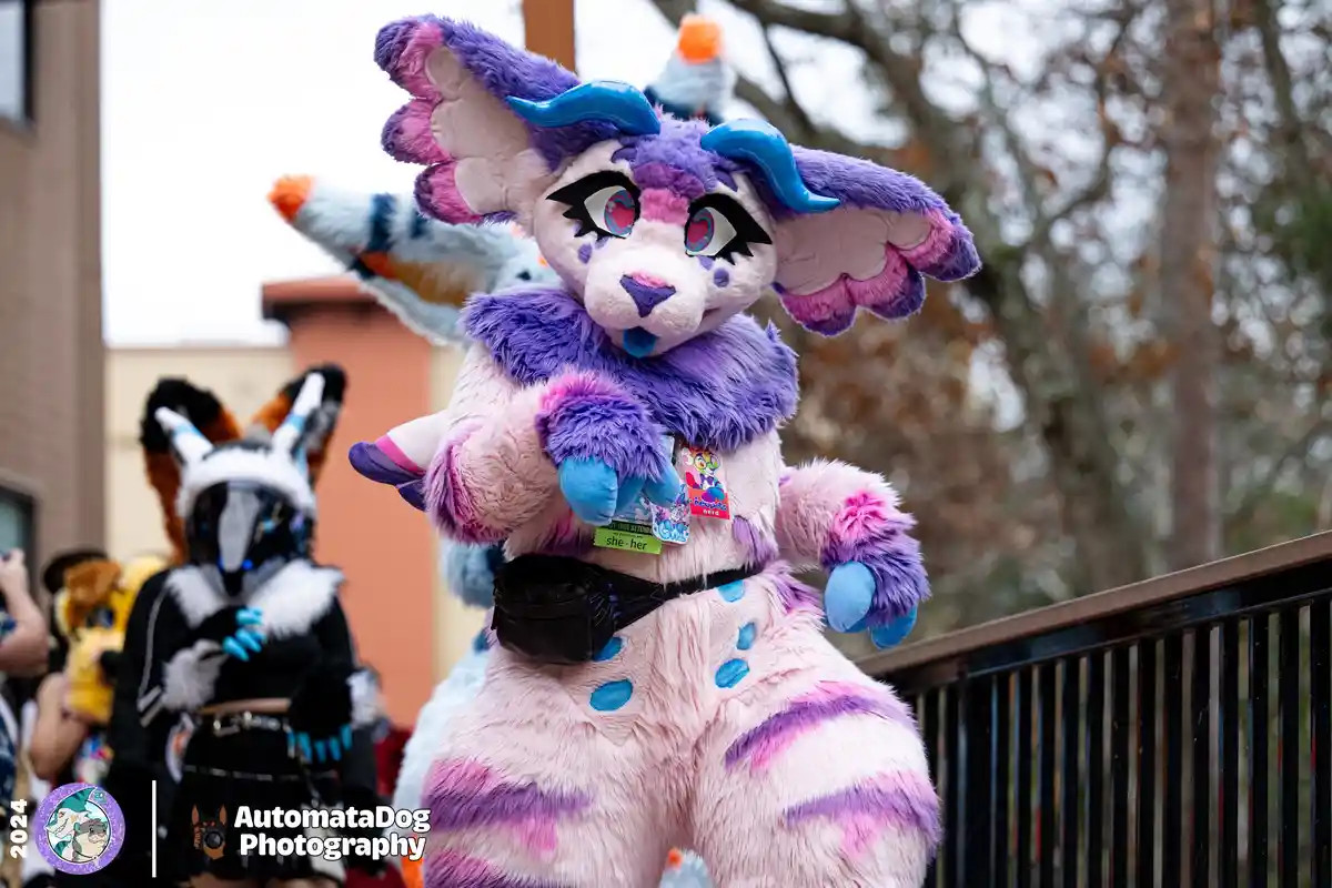 A person in a detailed pink, purple, and blue fursuit with large ears and horns poses outdoors, wearing a black waist pouch…