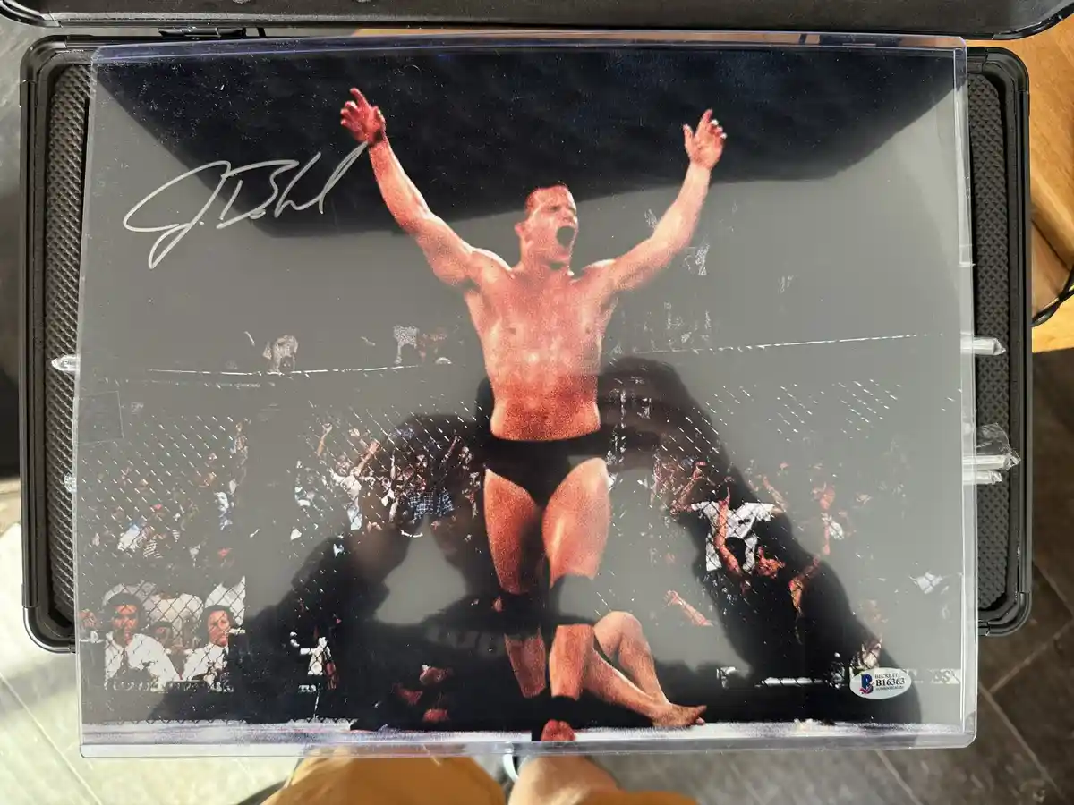 A signed, authenticated photograph of Georges St-Pierre celebrating a victory in a mixed martial arts cage, with his arms…