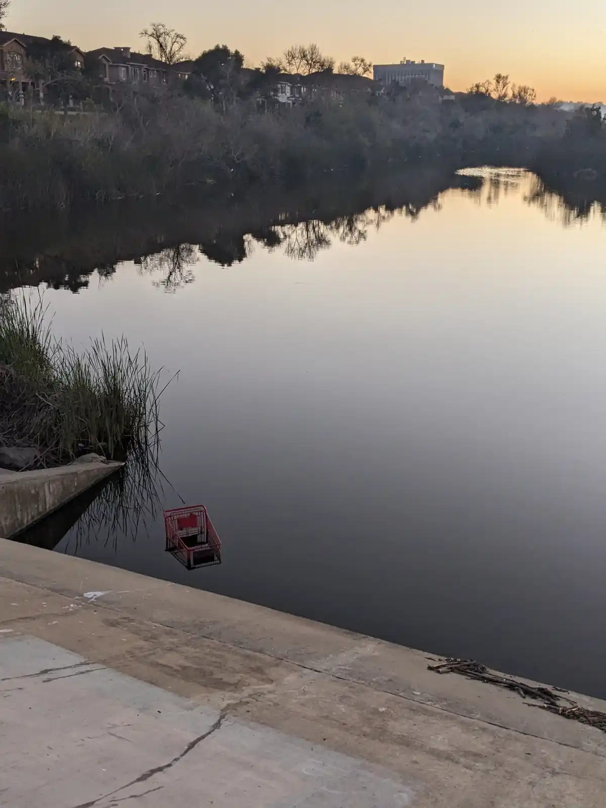 A red shopping cart is partially submerged in calm water at a lakeside, with a concrete walkway in the foreground and…