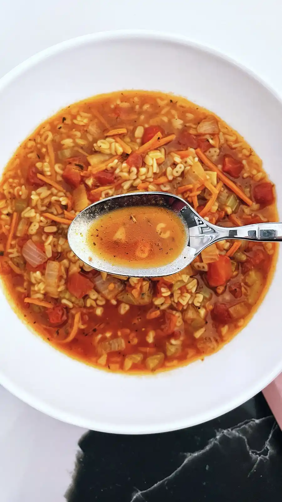 A close-up photo shows a white bowl filled with vegetable soup containing pasta, carrots, and tomatoes. A metal spoon is…