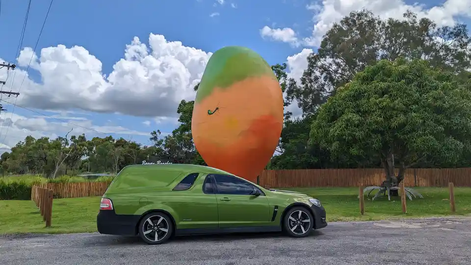 A lime-green station wagon is parked on a gravel lot in front of a giant, oversized mango sculpture painted with green and…