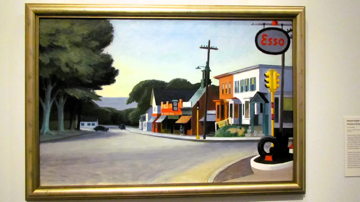 A framed painting of a quiet American street corner with colorful buildings, an Esso gas station sign, and a traffic light…
