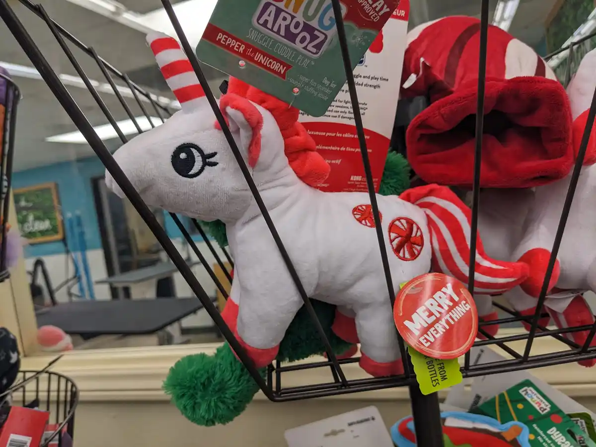 A white plush unicorn dog toy with red and white striped horn and tail, green pom-pom tail, and red circular patterns on its…