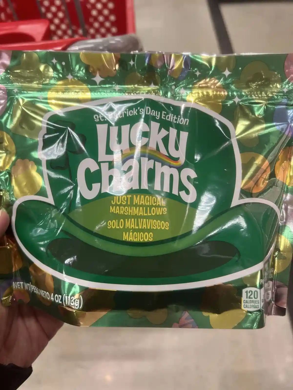 A hand holds a shiny green bag of Lucky Charms cereal, labeled 'St. Patrick's Day Edition' with a shamrock hat design. The…