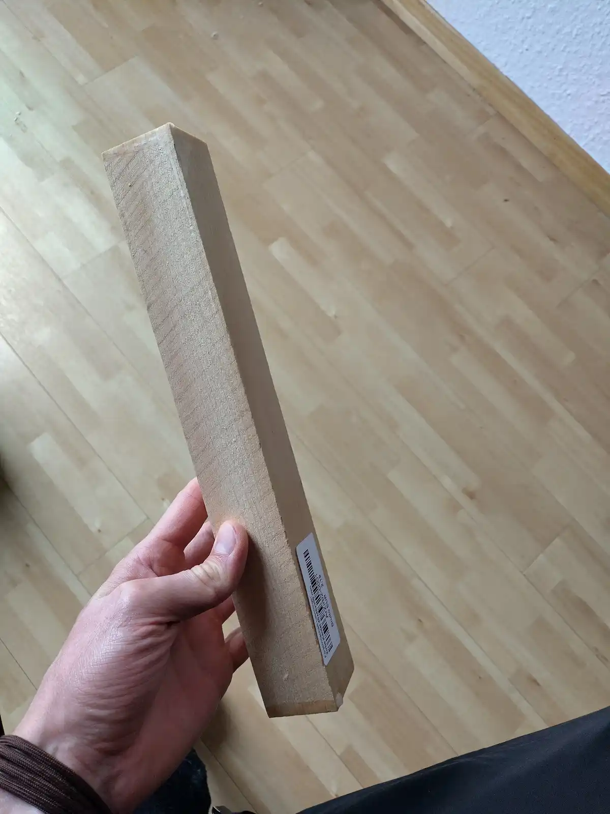 A hand holds a long, rectangular, light-colored wooden plank with a barcode sticker on its side, standing upright against a…