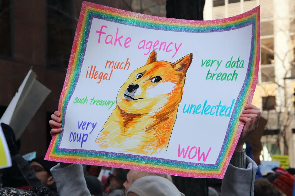 A person holds a handmade protest sign with a rainbow glitter border featuring a hand-drawn Shiba Inu dog. The sign has text…