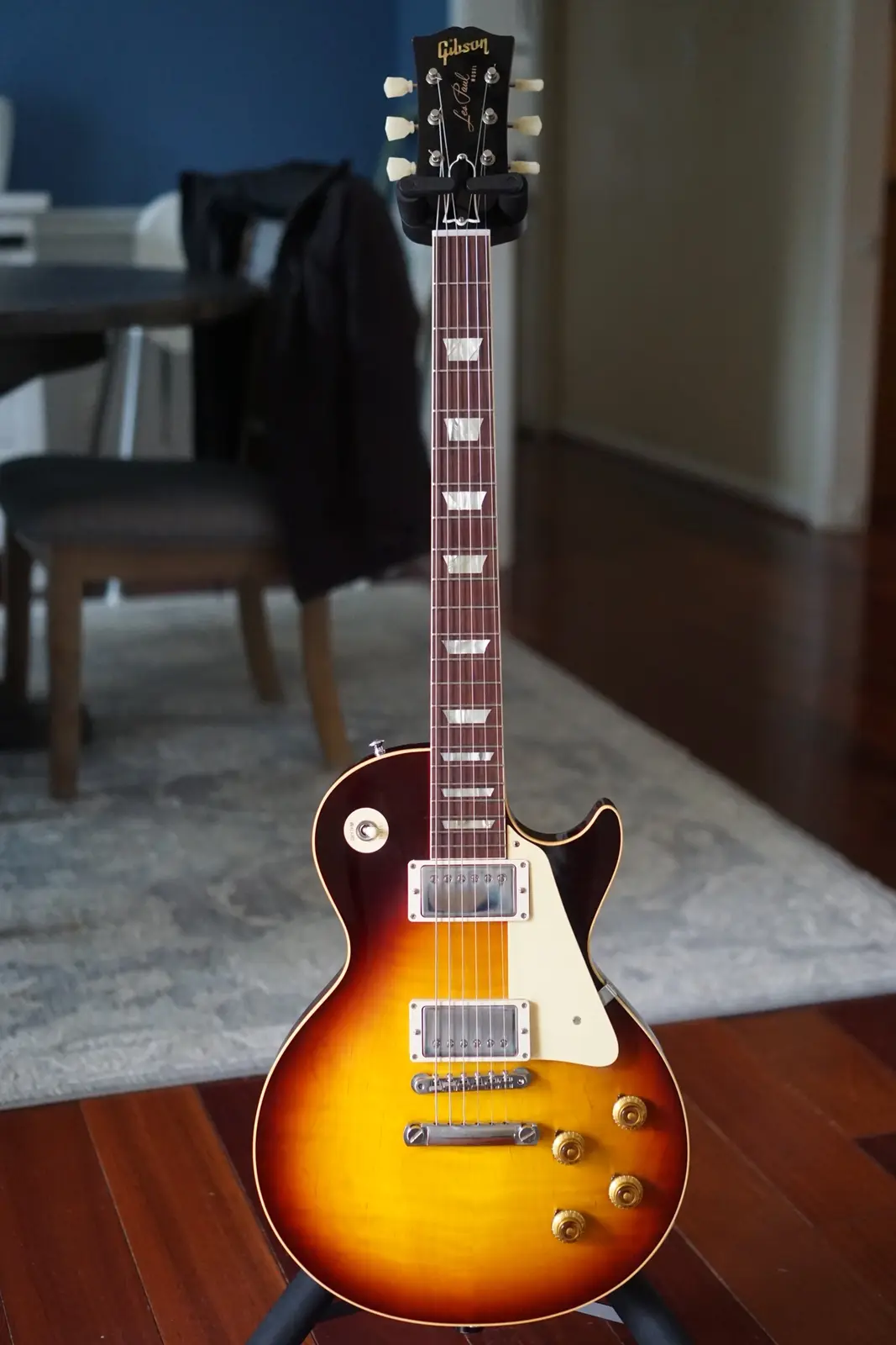 A Gibson Les Paul electric guitar with a sunburst finish and white pickguard is standing upright on a stand in a room with…