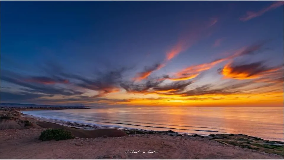 A stunning photograph of a beach at sunset, with vibrant orange and purple clouds over calm ocean waters, a sandy…