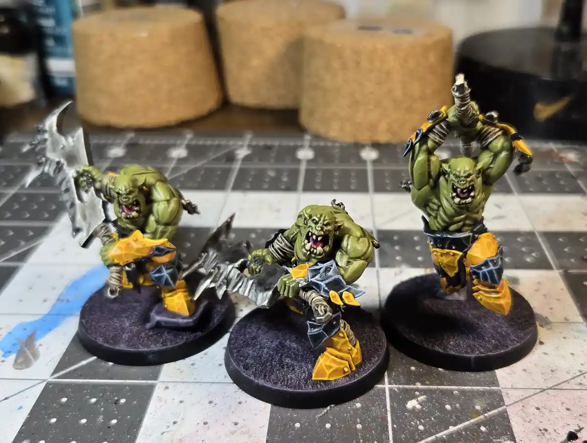 Three green, muscular orc miniatures with yellow and black armor stand on circular bases on a checkered, paint-splattered…