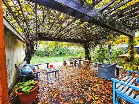A wooden deck covered in fallen autumn leaves, surrounded by garden furniture including benches, chairs, and tables, under a…