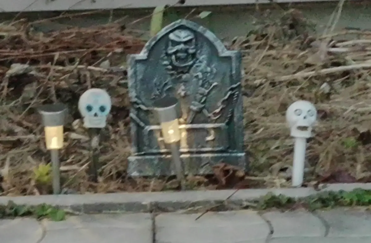 A blurry outdoor photo shows a small, dark-colored tombstone with a grim reaper figure on it, flanked by two small plastic…