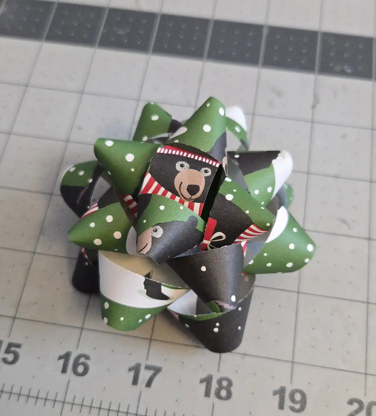 A handmade paper bow with green, black, and white sections featuring cartoon bears wearing red-and-white striped scarves…