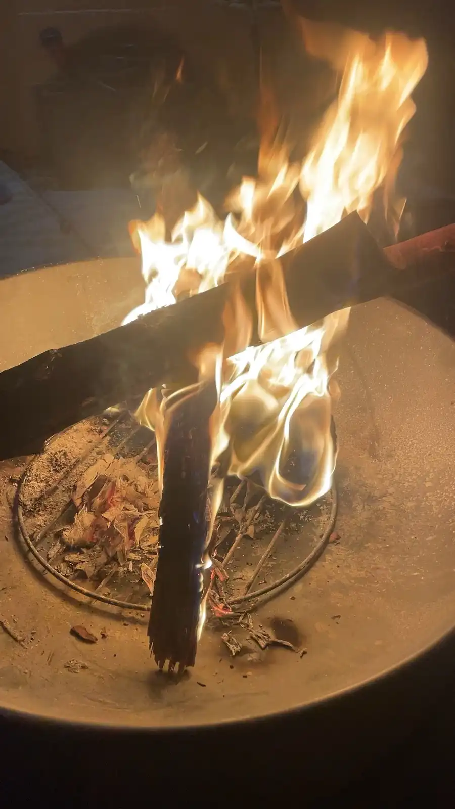 A close-up photo shows a large knife with a wooden handle burning intensely in a metal fire pit, flames licking up its…