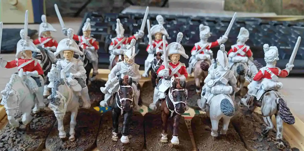 A row of painted miniature British cavalry soldiers from the 18th century, mostly in red coats and white helmets, are…