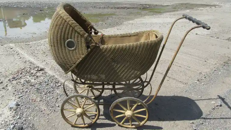 An old-fashioned wicker baby pram with wooden wheels and a metal frame sits abandoned on a gravelly, muddy ground near a…