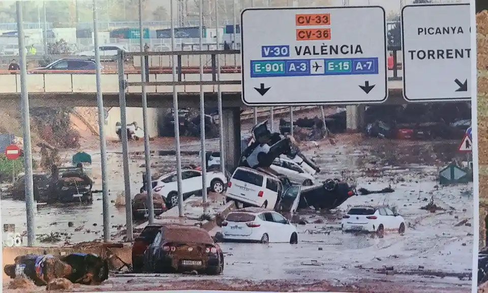 A photo shows a flooded and debris-strewn highway underpass with many cars submerged or overturned in muddy water. Above the…