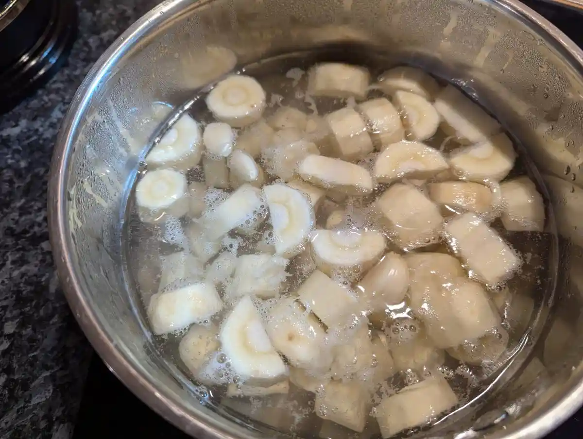 A stainless steel pot on a stovetop contains water with chunks of peeled, sliced bananas boiling, creating bubbles and…