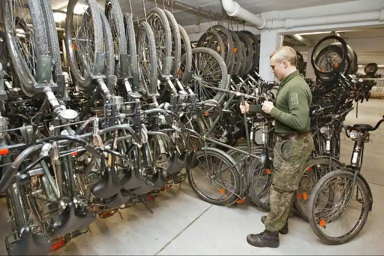 A soldier in military fatigues stands in a warehouse surrounded by rows of disassembled bicycles, mostly missing wheels and…