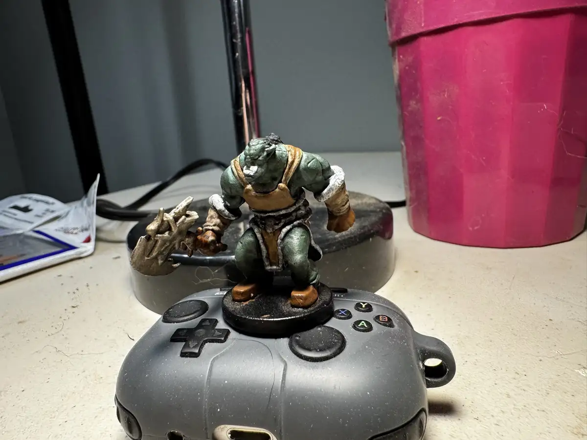 A painted green orc miniature figurine, holding a bone-like weapon, is placed on top of a grey Xbox controller. The…